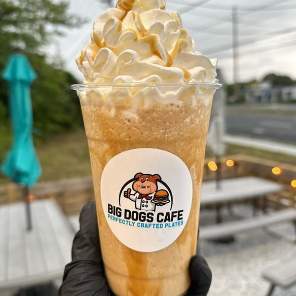 Hand holding a Big Dogs Cafe caramel frappe with whipped cream and caramel drizzle outdoors.