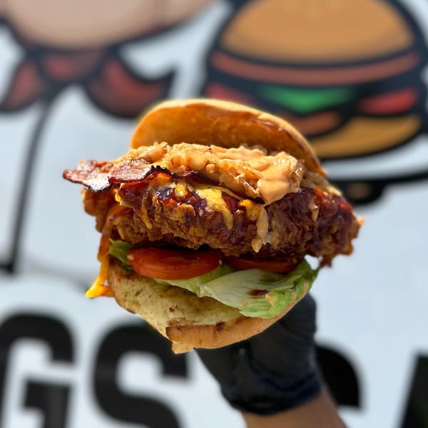 A Big Dogs fried chicken sandwich with bacon, cheese, lettuce, and tomato, held by a gloved hand.